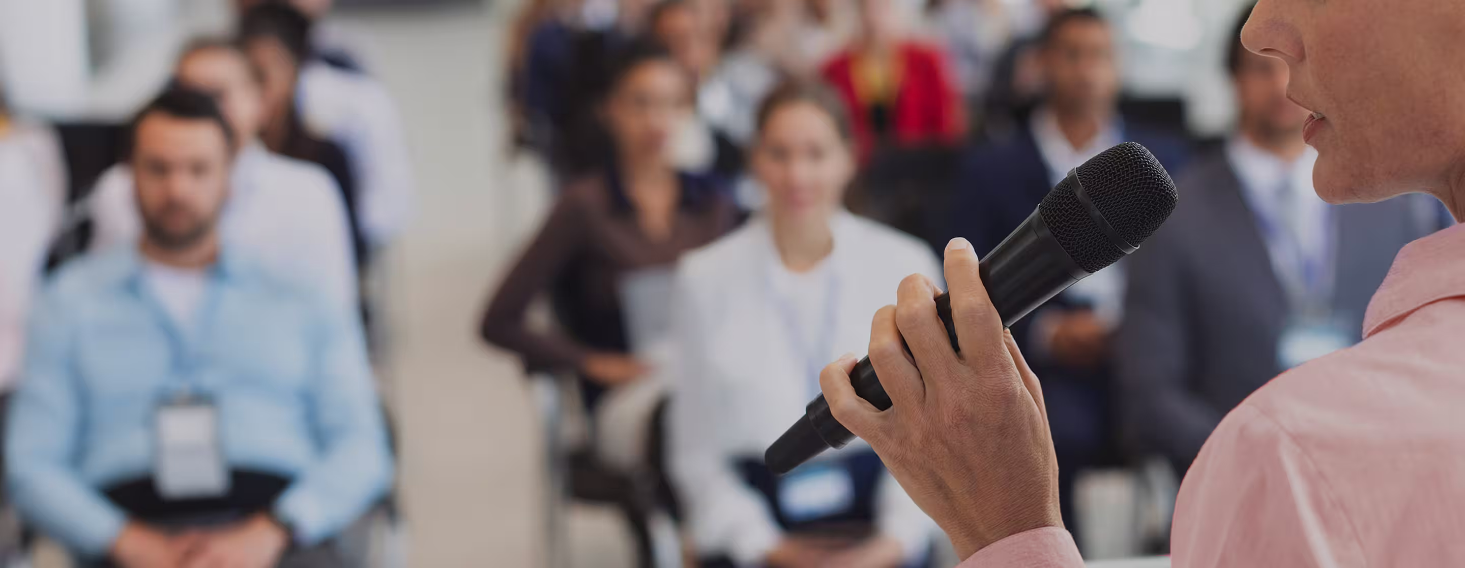 Close-up of a person holding a microphone speaking to a blurred audience sitting and listening.