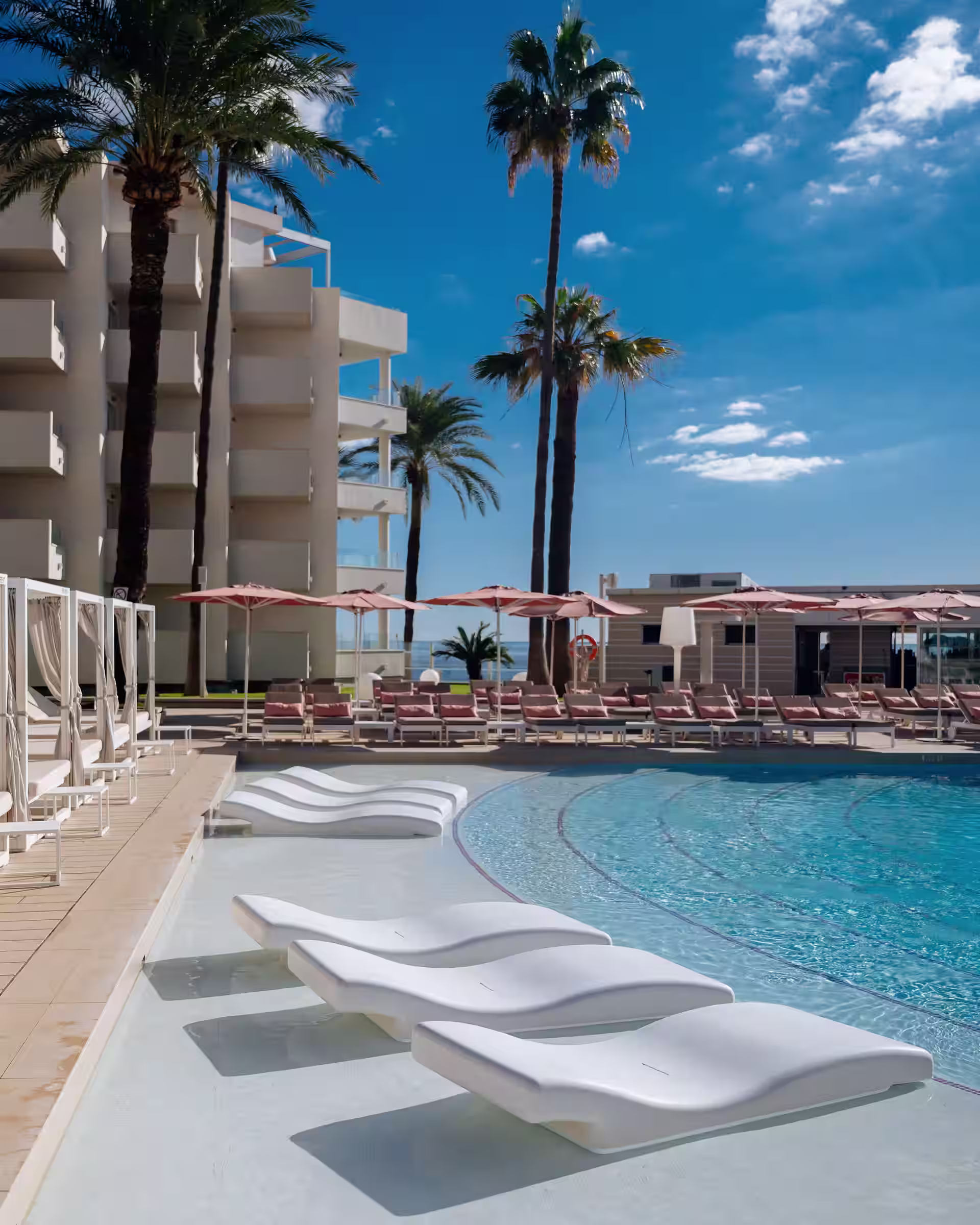 White modern lounge chairs in shallow pool area with palm trees and sunbeds under umbrellas nearby.