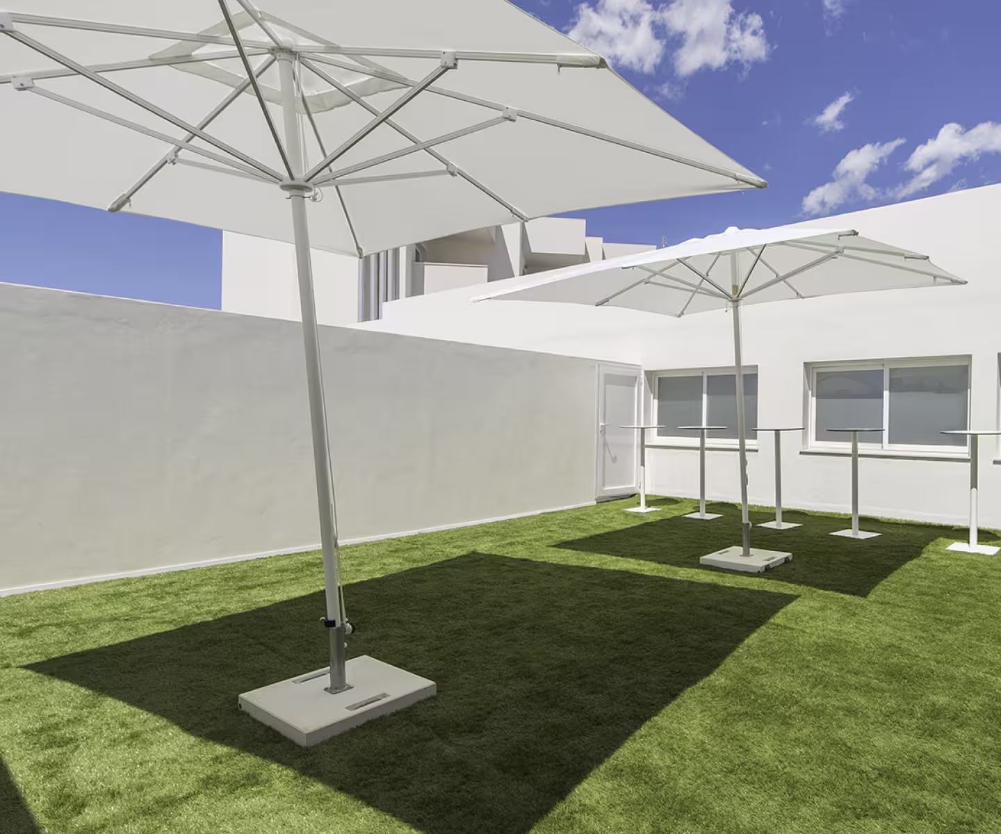 Two large white patio umbrellas casting shadows on green artificial grass in a white-walled courtyard.
