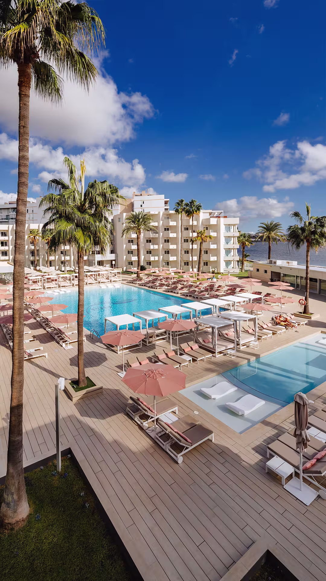 Resort swimming pool area with lounge chairs, pink umbrellas, palm trees, and a hotel building under a sunny blue sky.