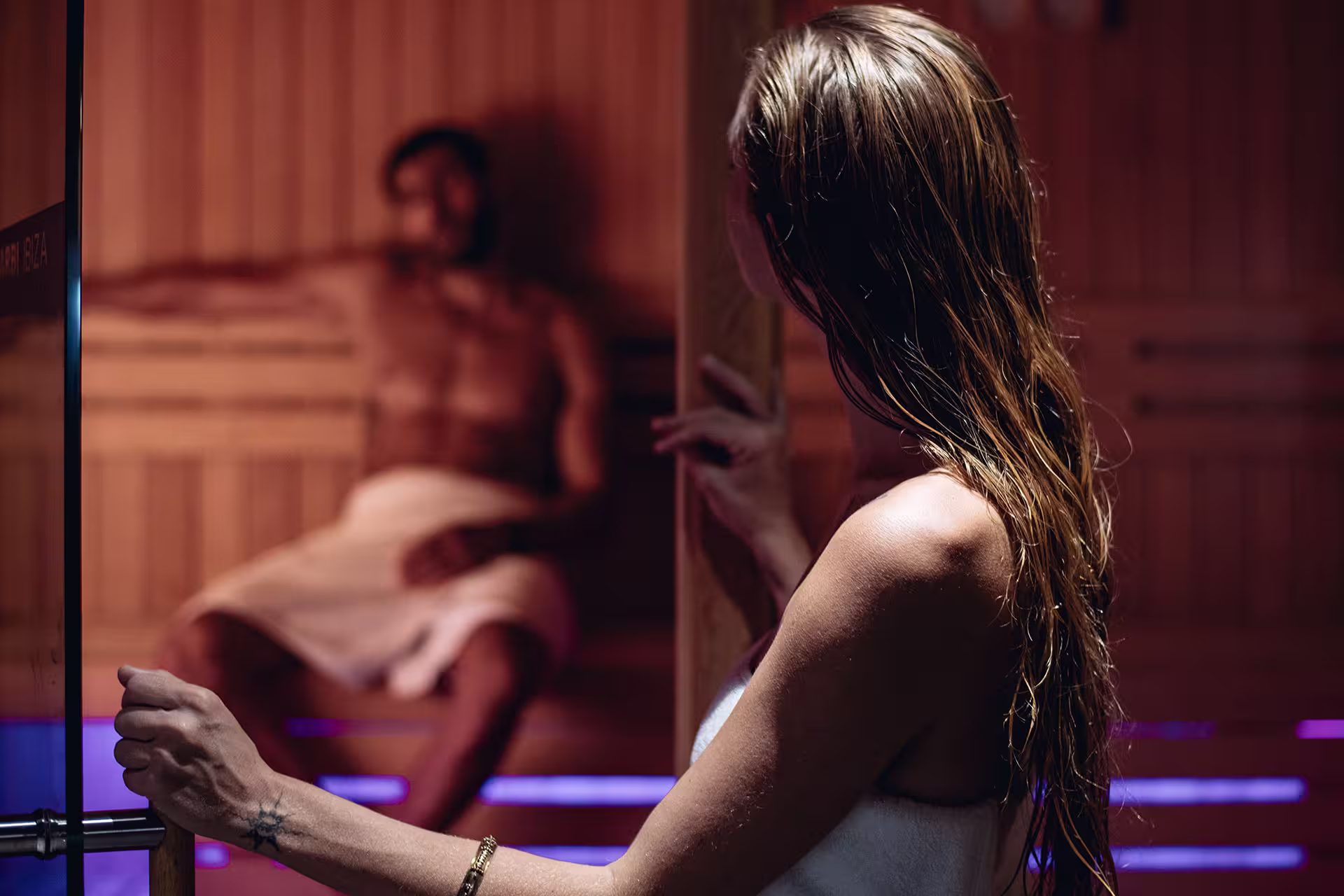 Woman entering a sauna where a man wrapped in a towel is sitting on a wooden bench.