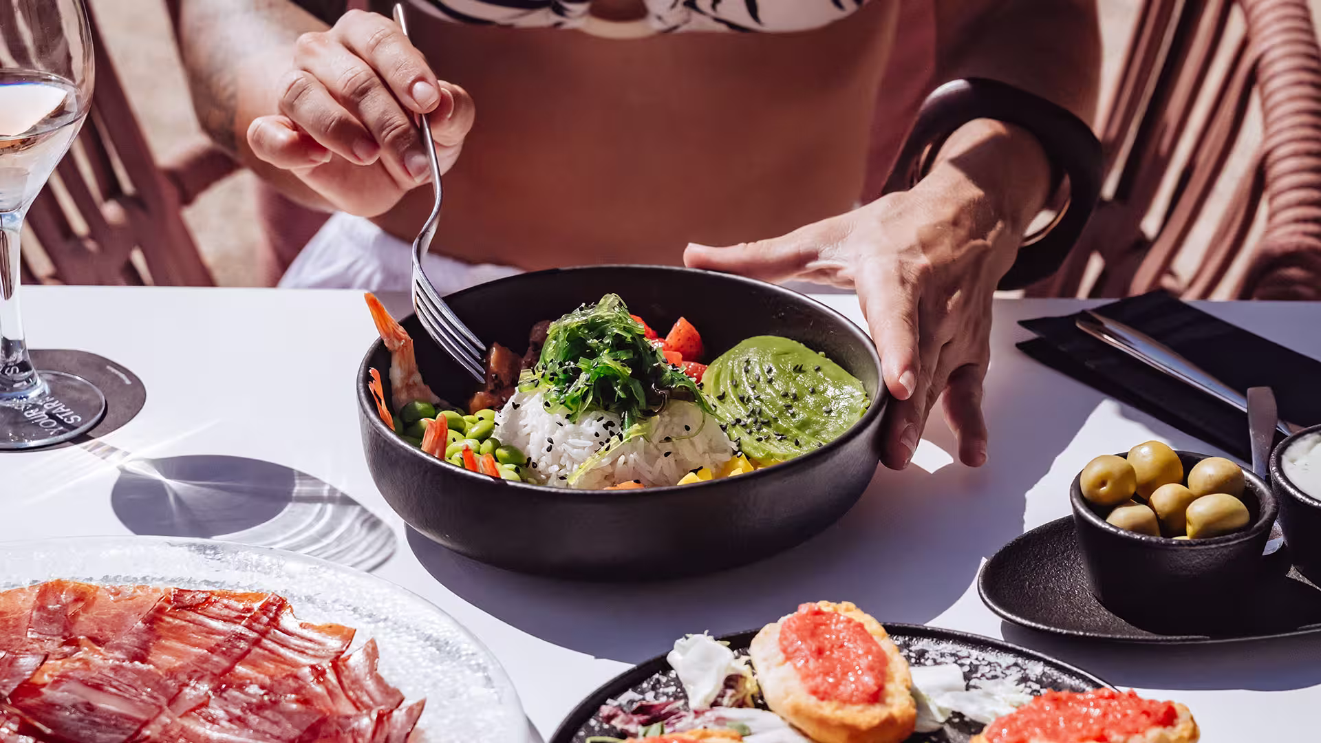 Person eating a colorful poke bowl with shrimp, avocado, rice, and vegetables at a sunny outdoor table.