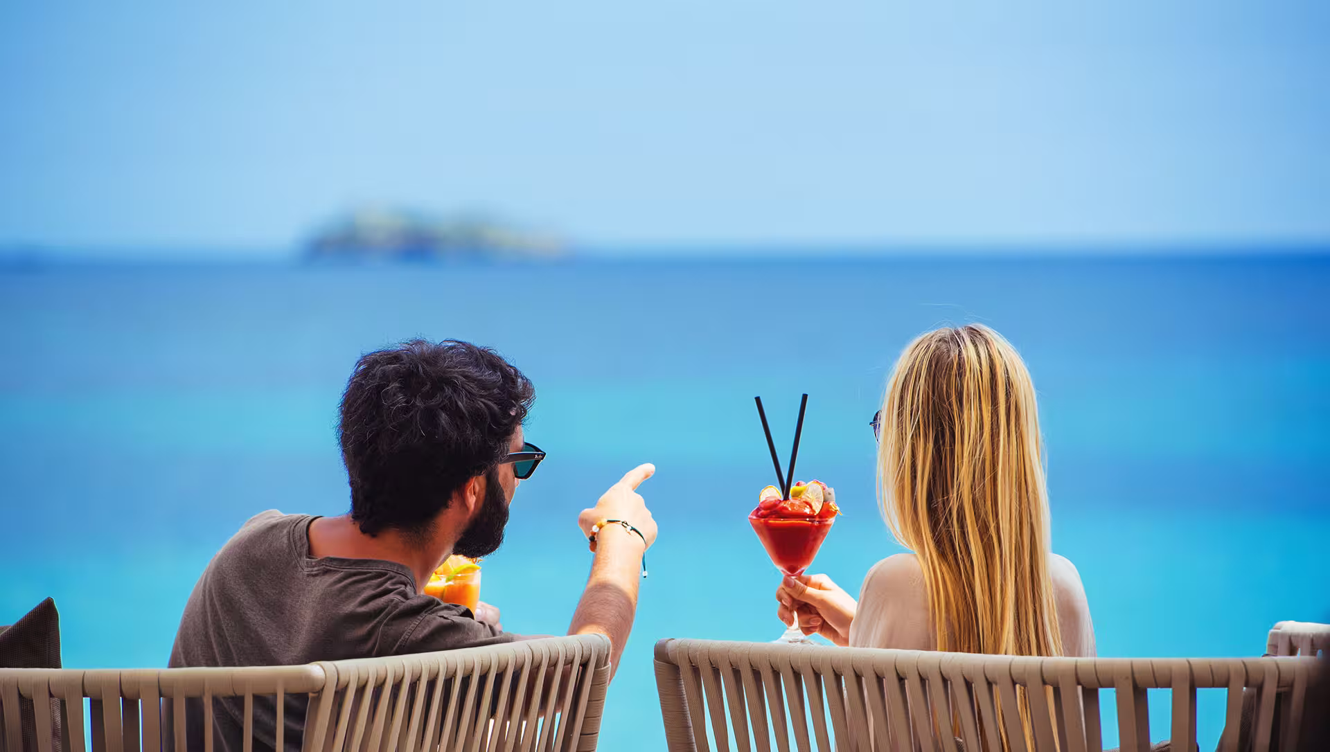 Man and woman sitting on chairs facing the ocean, holding colorful cocktails with straws.