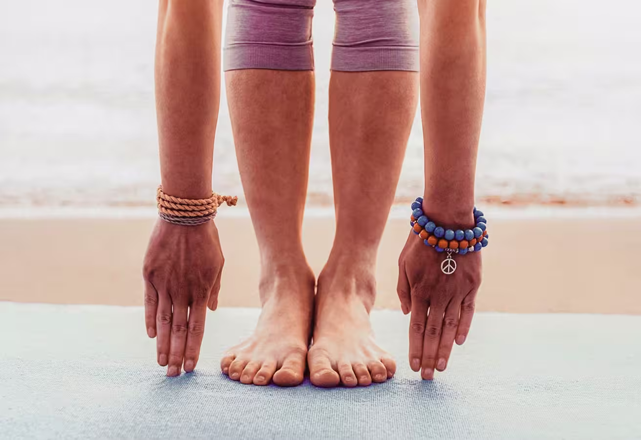 Person standing barefoot on a mat by the beach, bending forward to touch the ground with both hands wearing beaded bracelets.