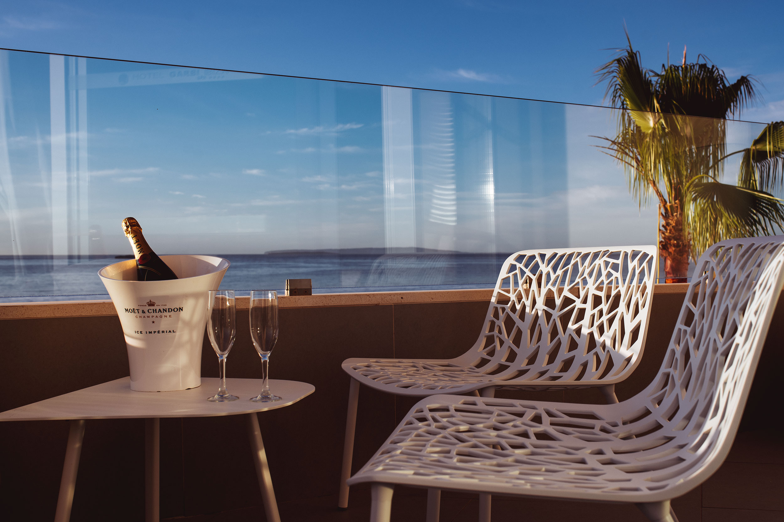 Two empty champagne flutes beside a Moët & Chandon champagne bottle in an ice bucket on a table, with white modern chairs and a sea view in the background.
