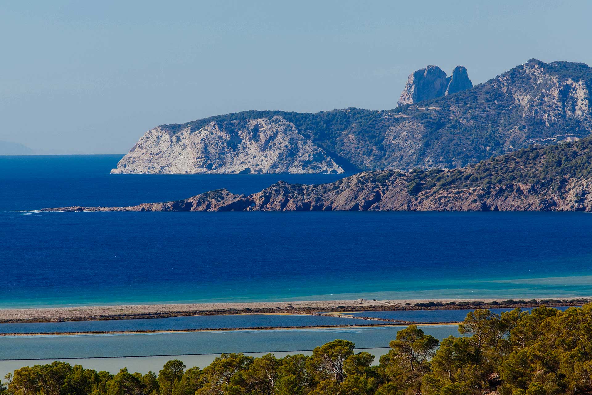 Coastal landscape showing layered blue sea waters, rocky hills, and green trees under a clear sky.