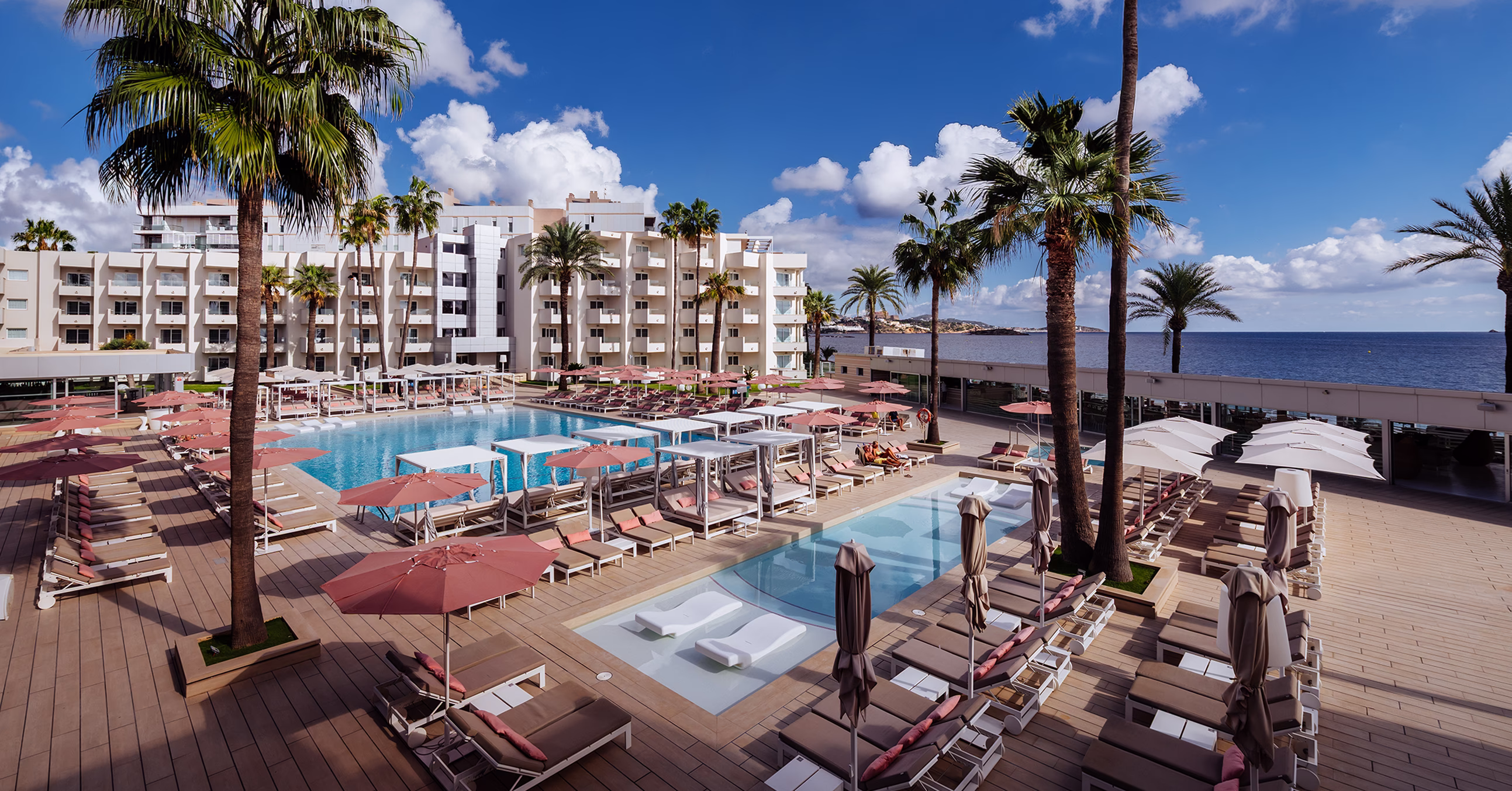 Resort swimming pool area with lounge chairs, pink umbrellas, palm trees, and a hotel building under a sunny blue sky.