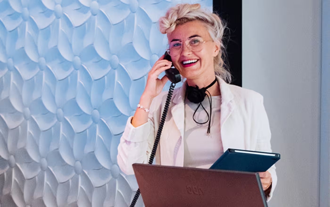 Smiling woman with glasses talking on a corded phone while holding a tablet in an office.