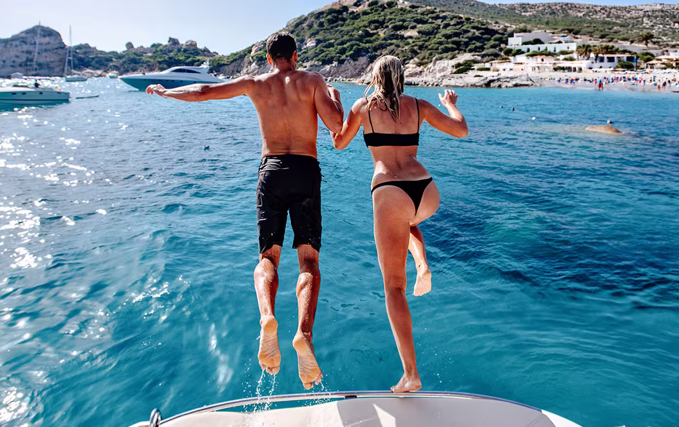 Couple holding hands jumping off a boat into clear blue sea near rocky coastline.