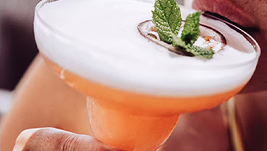 Close-up of a person sipping a frothy orange cocktail garnished with mint leaves.