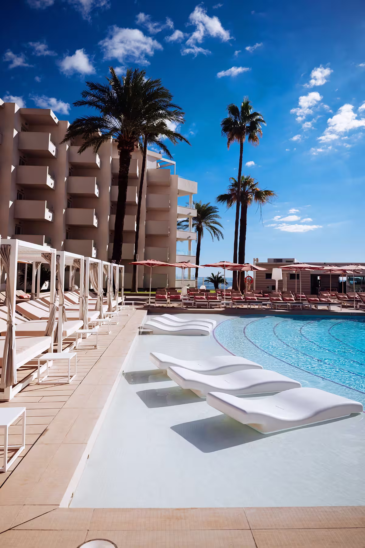 Modern outdoor pool area with white lounge chairs partially submerged in water, surrounded by palm trees and a building under a blue sky.