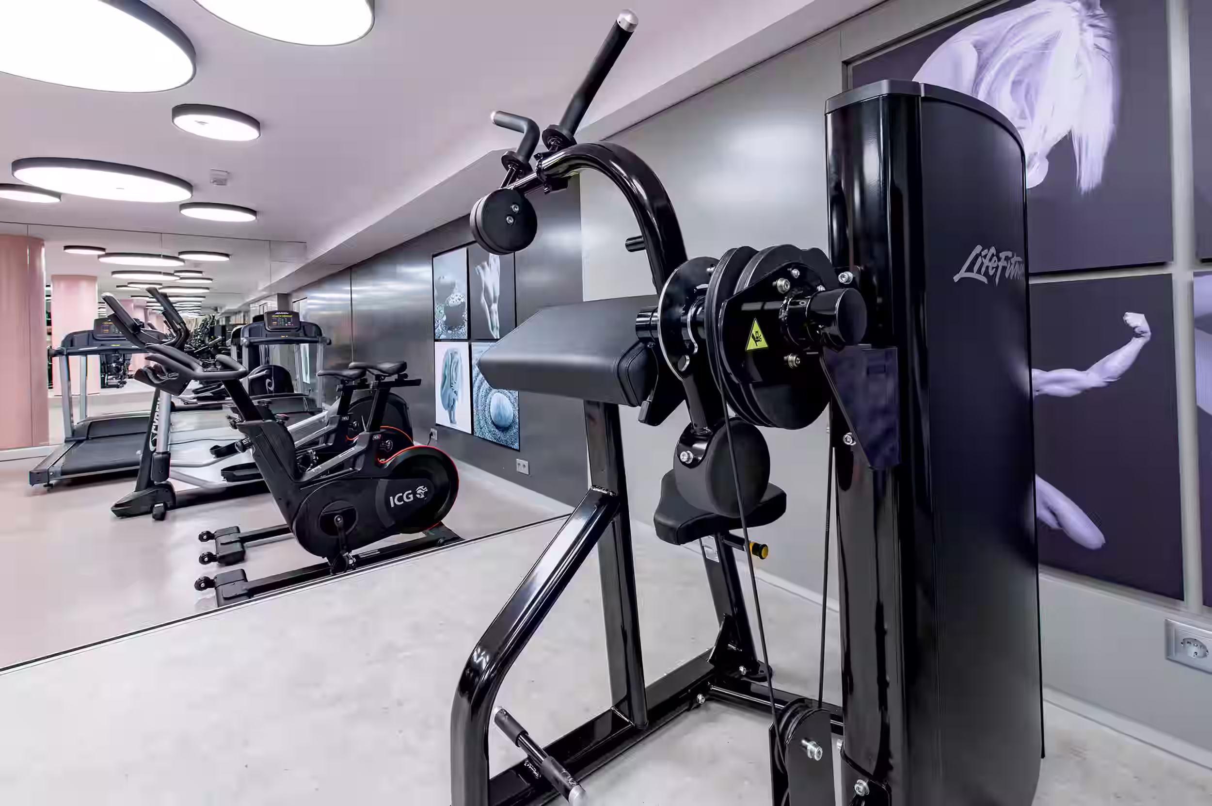 Modern gym with black adjustable benches, dumbbells, weight machines, pink pillars, and circular ceiling lights.