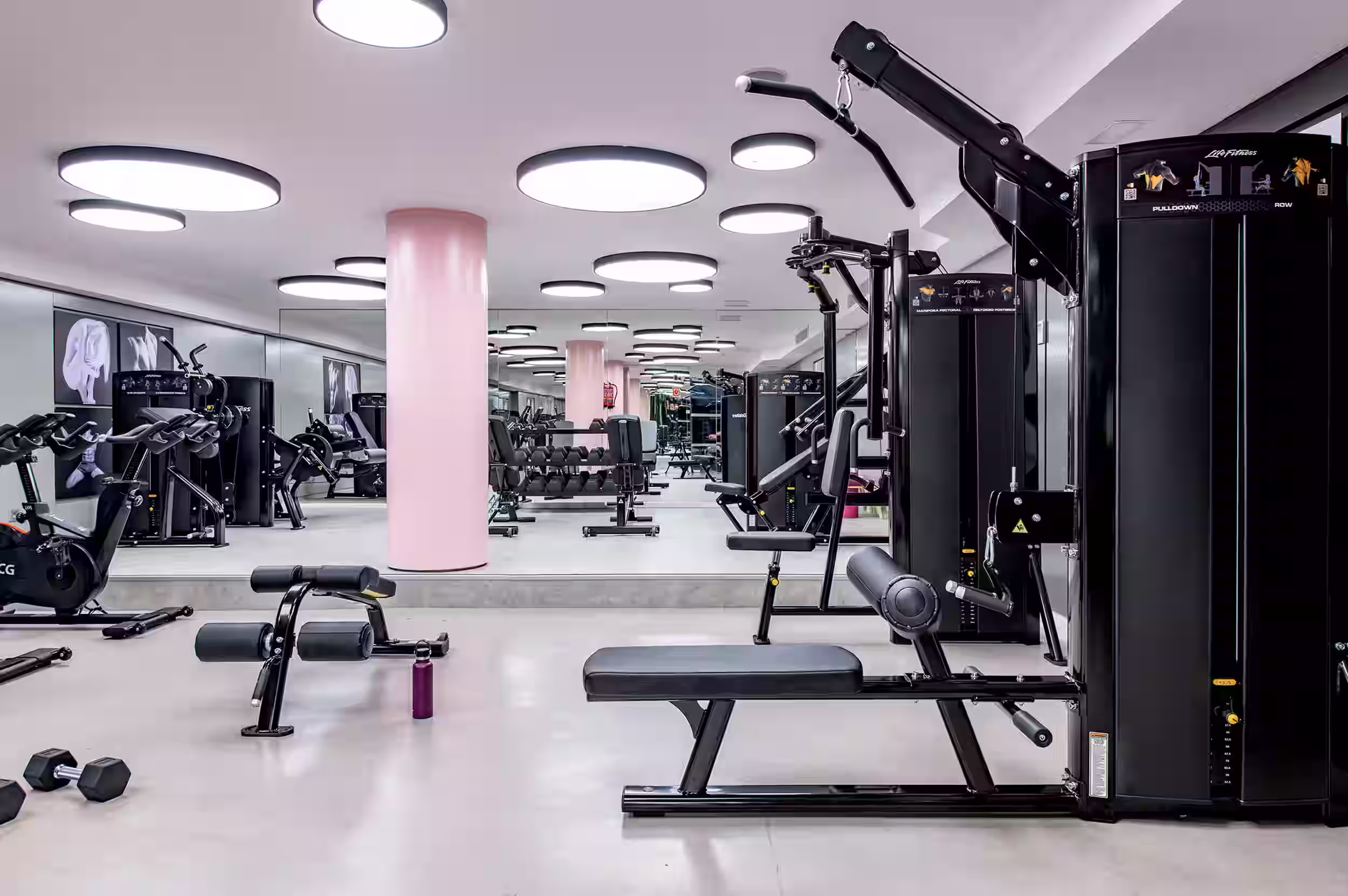 Modern gym interior with weight machines, exercise bike, dumbbells, and circular ceiling lights.
