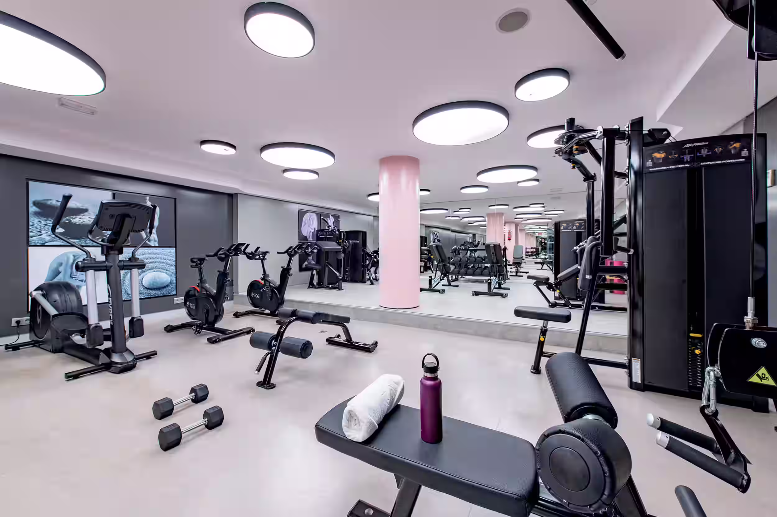Modern gym with black adjustable benches, dumbbells, weight machines, pink pillars, and circular ceiling lights.