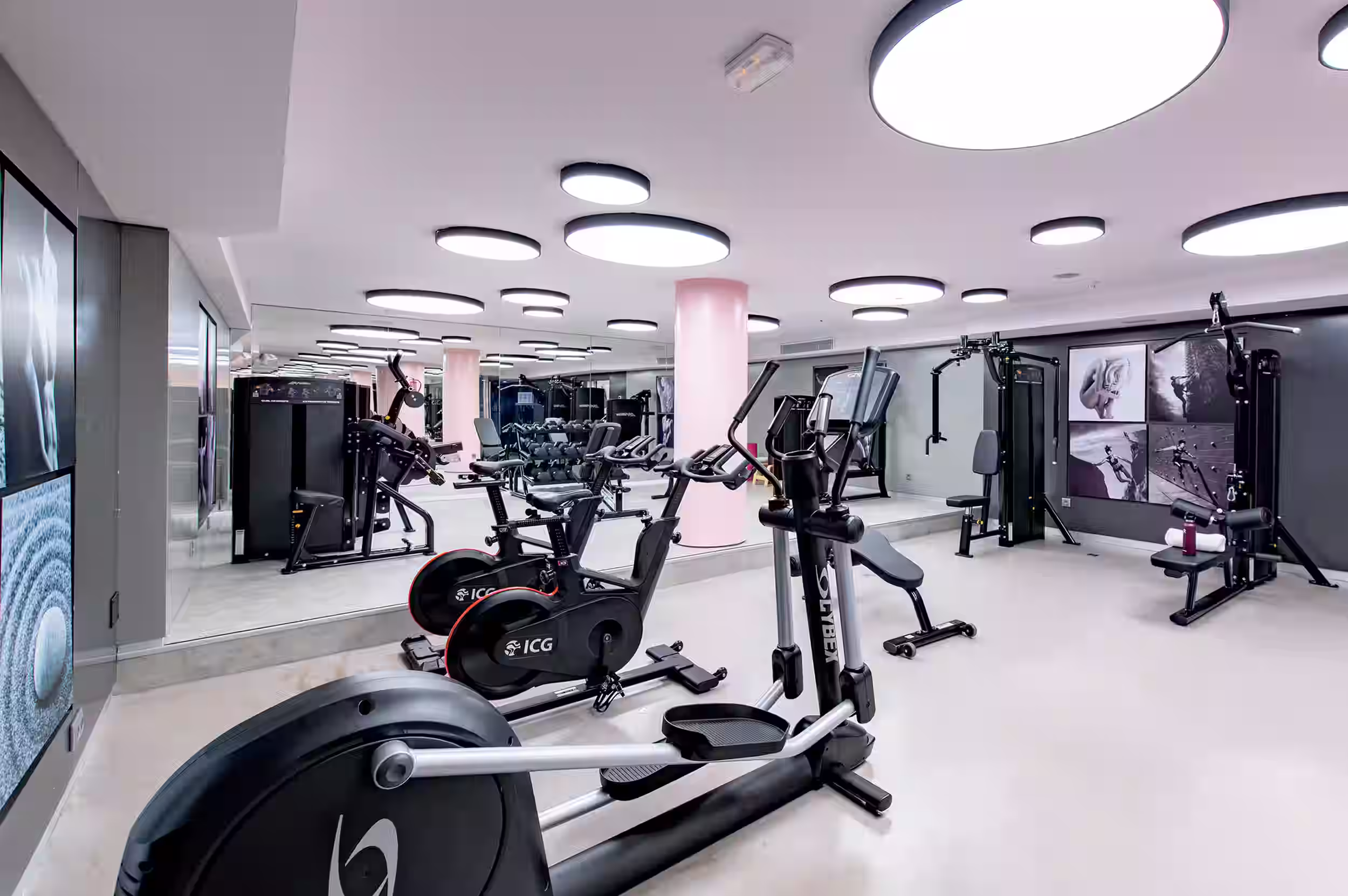 Modern gym with black adjustable benches, dumbbells, weight machines, pink pillars, and circular ceiling lights.
