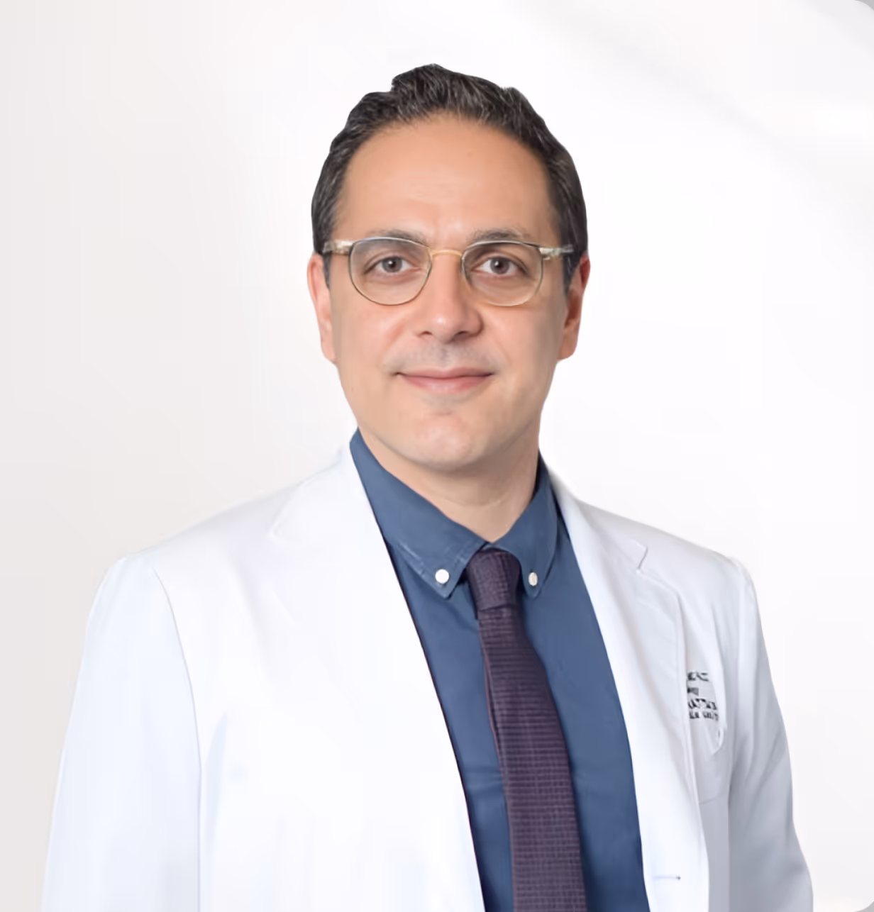 Portrait of a man wearing glasses, a white medical coat, blue shirt, and dark tie against a white background.