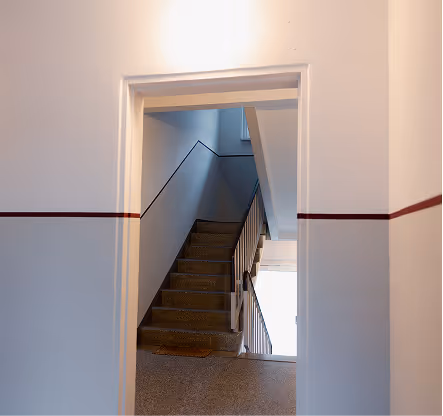 Blick durch eine Tür auf eine Treppe in einem weißen Treppenhaus mit braunem Streifen an den Wänden.
