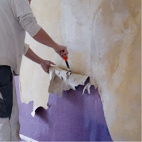 Person entfernt Tapete mit einem Messer, darunter ist eine violette Wand sichtbar.