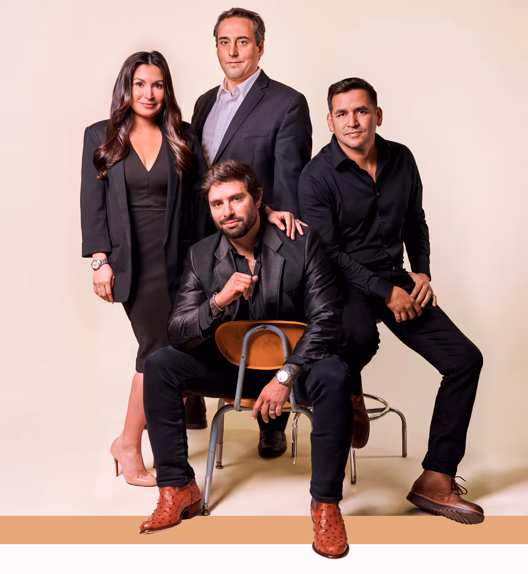 Four professionally dressed individuals posing together against a beige background, three standing and one sitting backward on a chair.