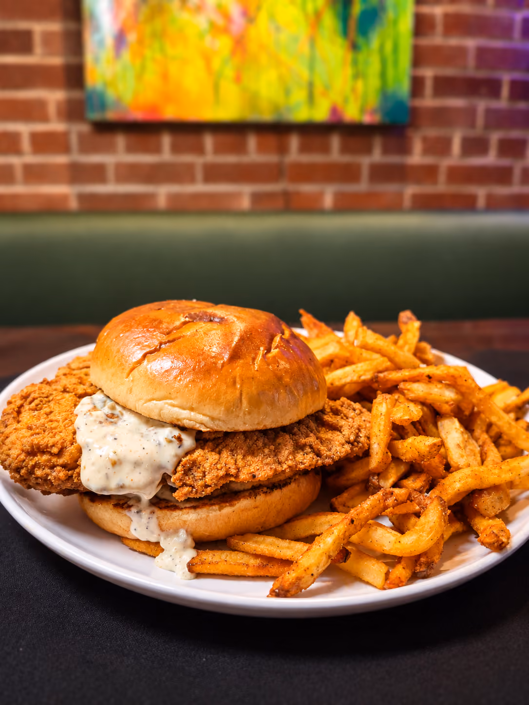 Plate with a crispy fried chicken sandwich topped with white gravy and a lightly toasted bun, served with seasoned French fries.