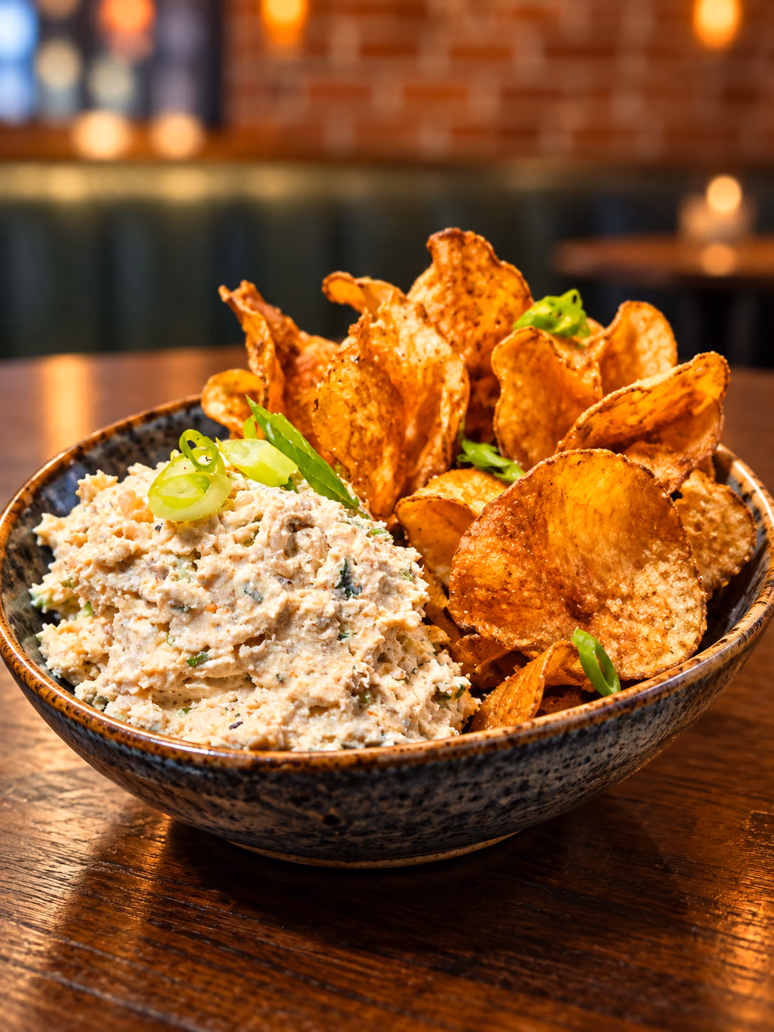 Bowl of seasoned potato chips served with a creamy dip garnished with sliced green onions on a wooden table with a blurred warm-toned background.