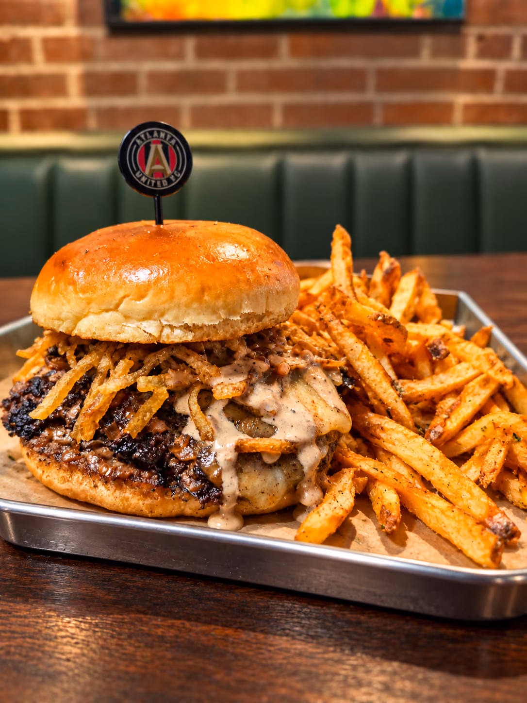 Close-up of a gourmet cheeseburger with crispy fried onion strings and creamy sauce on a toasted bun, served with a side of seasoned fries on a metal tray in a restaurant setting.