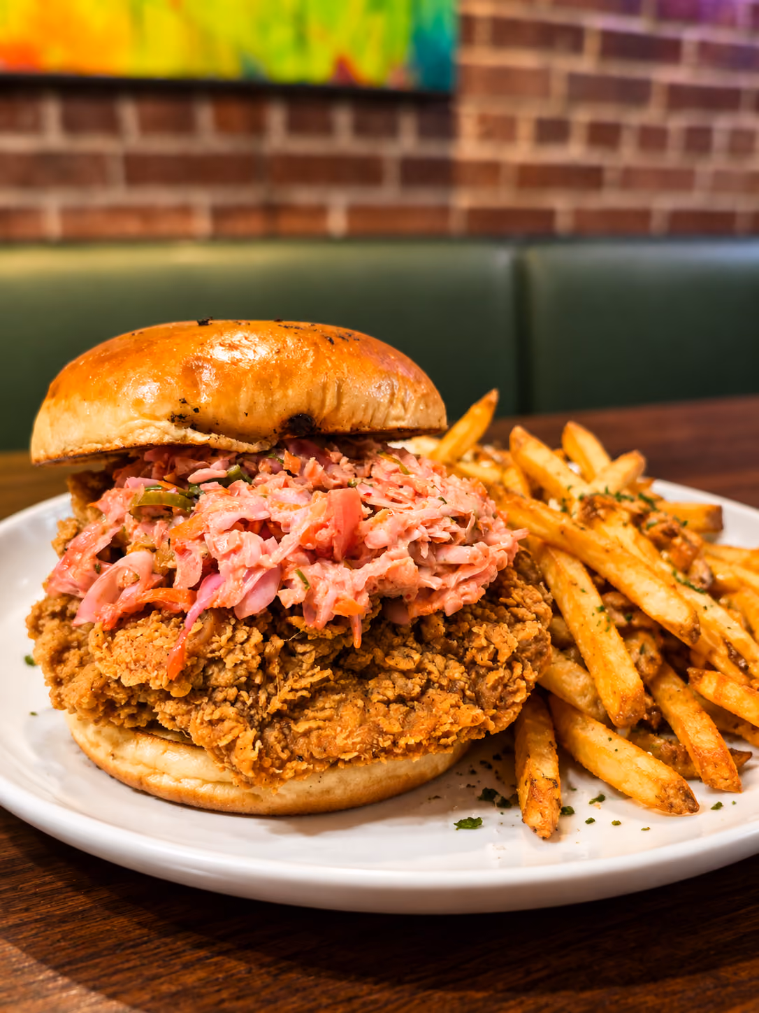 Fried chicken sandwich topped with shredded pickled vegetables on a toasted bun, served with seasoned French fries on a white plate.