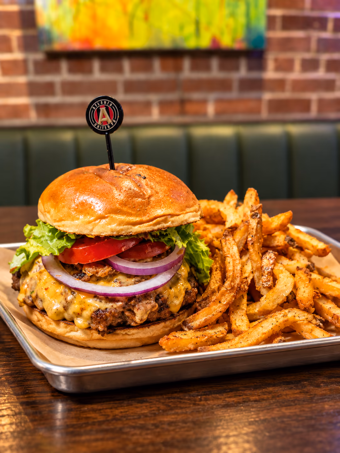Cheeseburger with lettuce, tomato, and red onion on a toasted bun served with seasoned French fries on a metal tray in a restaurant setting.