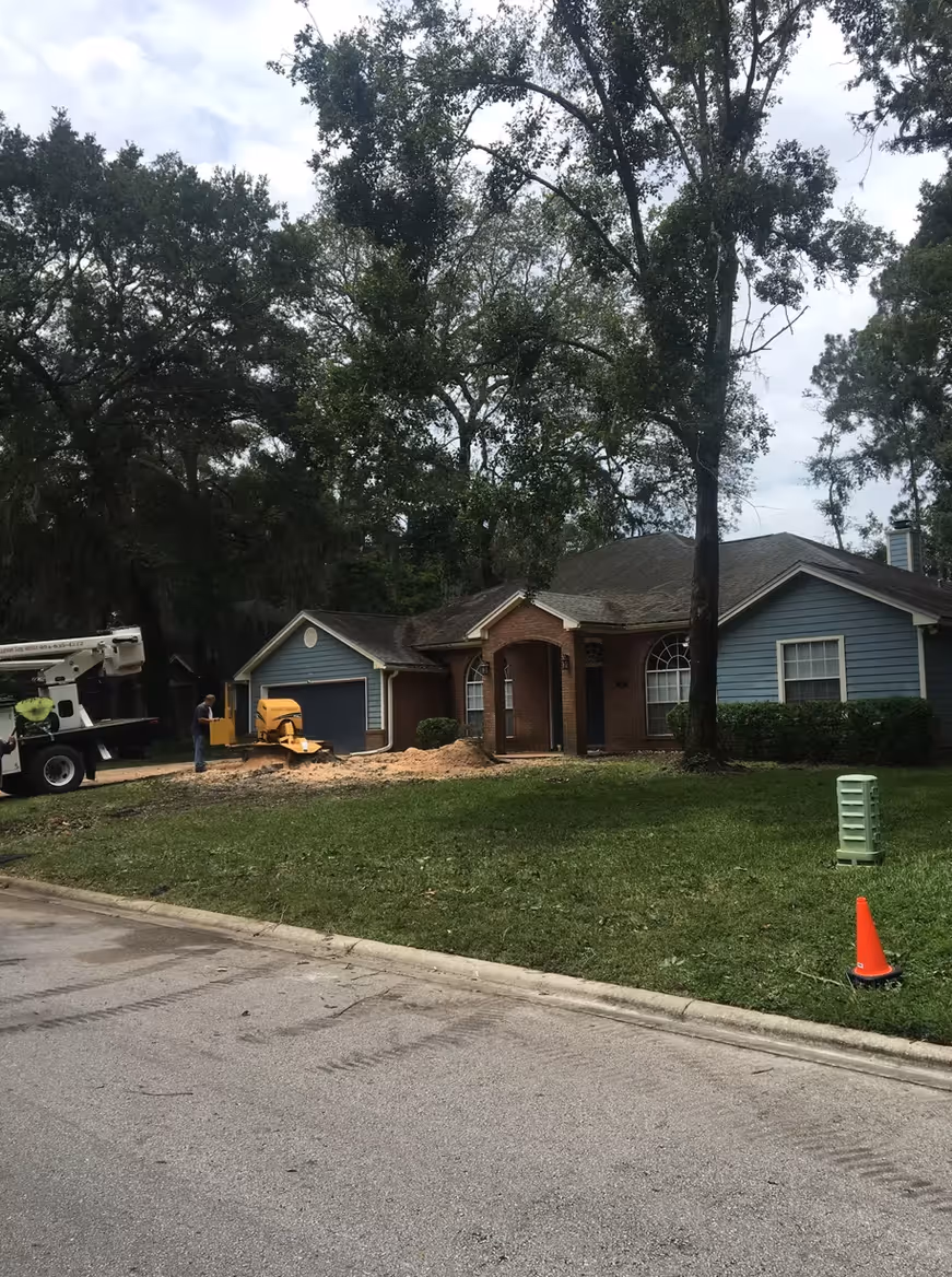 A worker uses a wood chipper on the front lawn of a suburban house with large trees and a blue garage door.