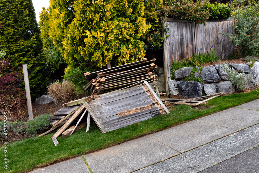 Disassembled old wooden fence pieces stacked on grass next to a concrete sidewalk with greenery and a wooden fence in the background.