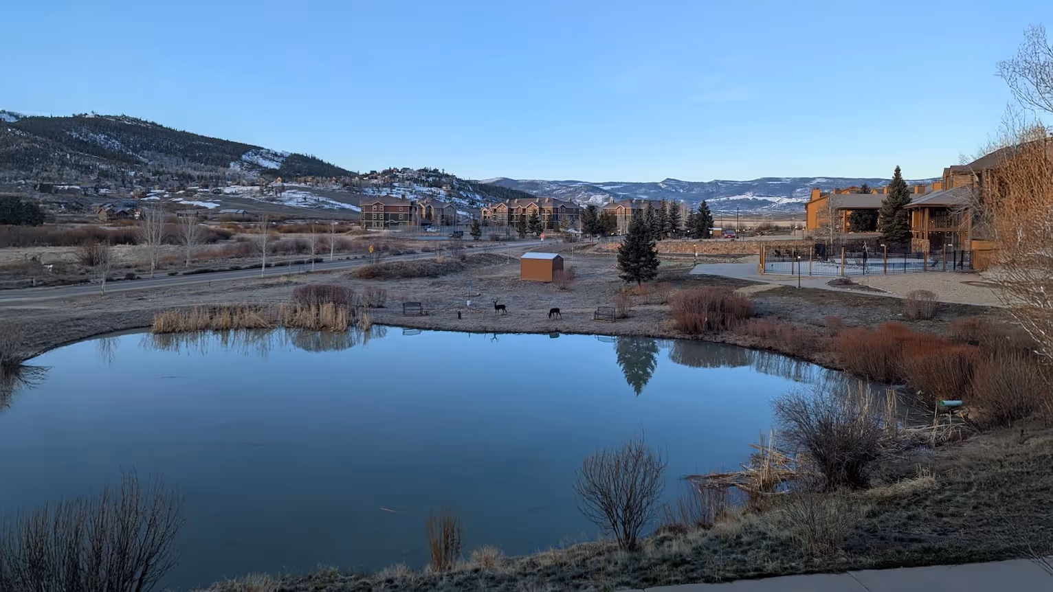 A serene landscape with a calm pond reflecting the clear blue sky. Surrounding the water are sparse trees, brown vegetation, and distant snow-capped hills.