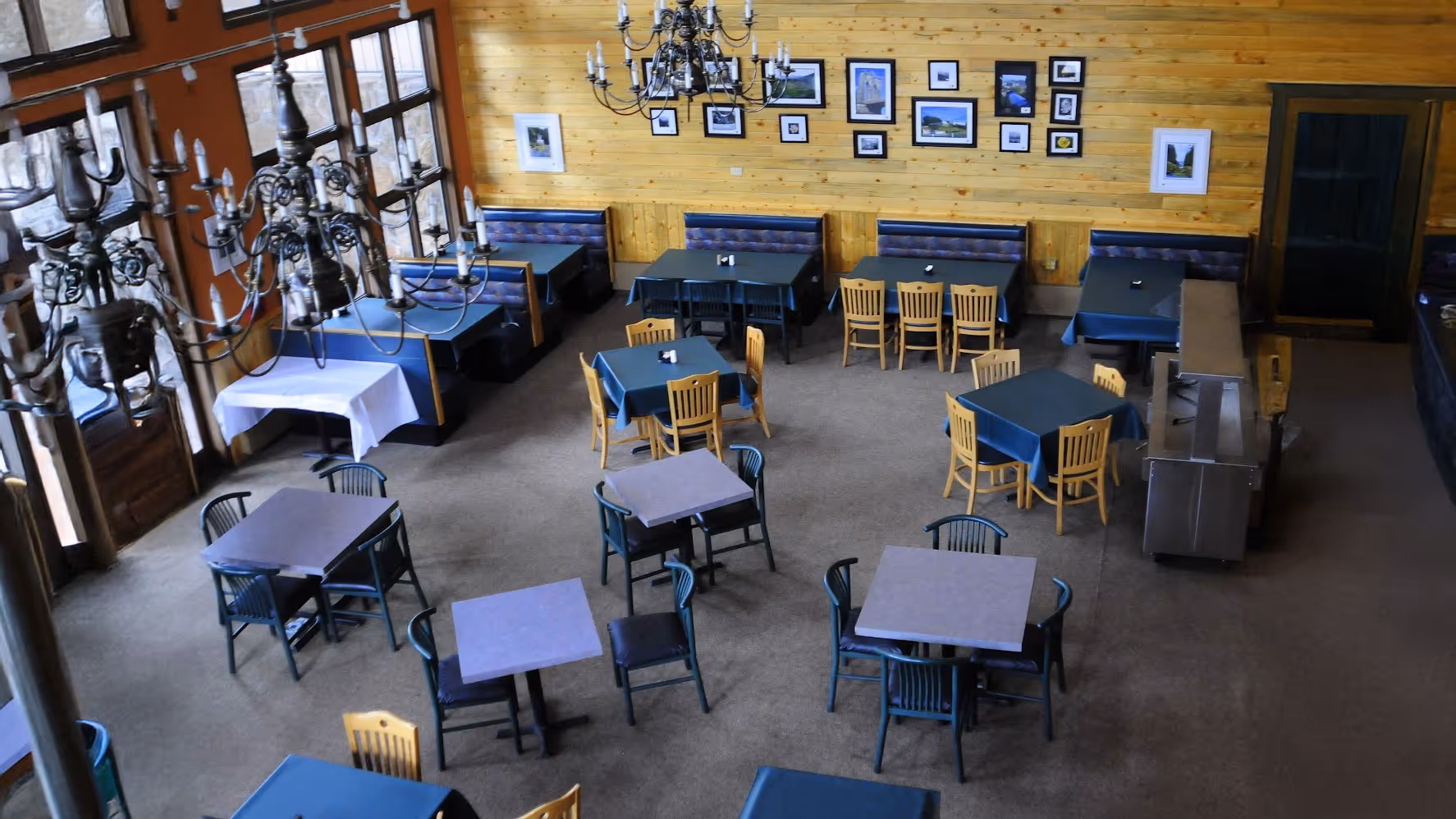 A spacious restaurant interior with wooden walls, adorned with framed photos. Empty tables and chairs are neatly arranged beneath elegant chandeliers, conveying a cozy yet formal ambiance.