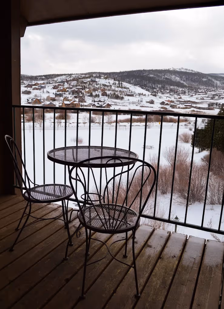 A wooden balcony features two wrought iron chairs and a table overlooking a snowy landscape with hills and scattered houses, conveying a serene, wintery setting.