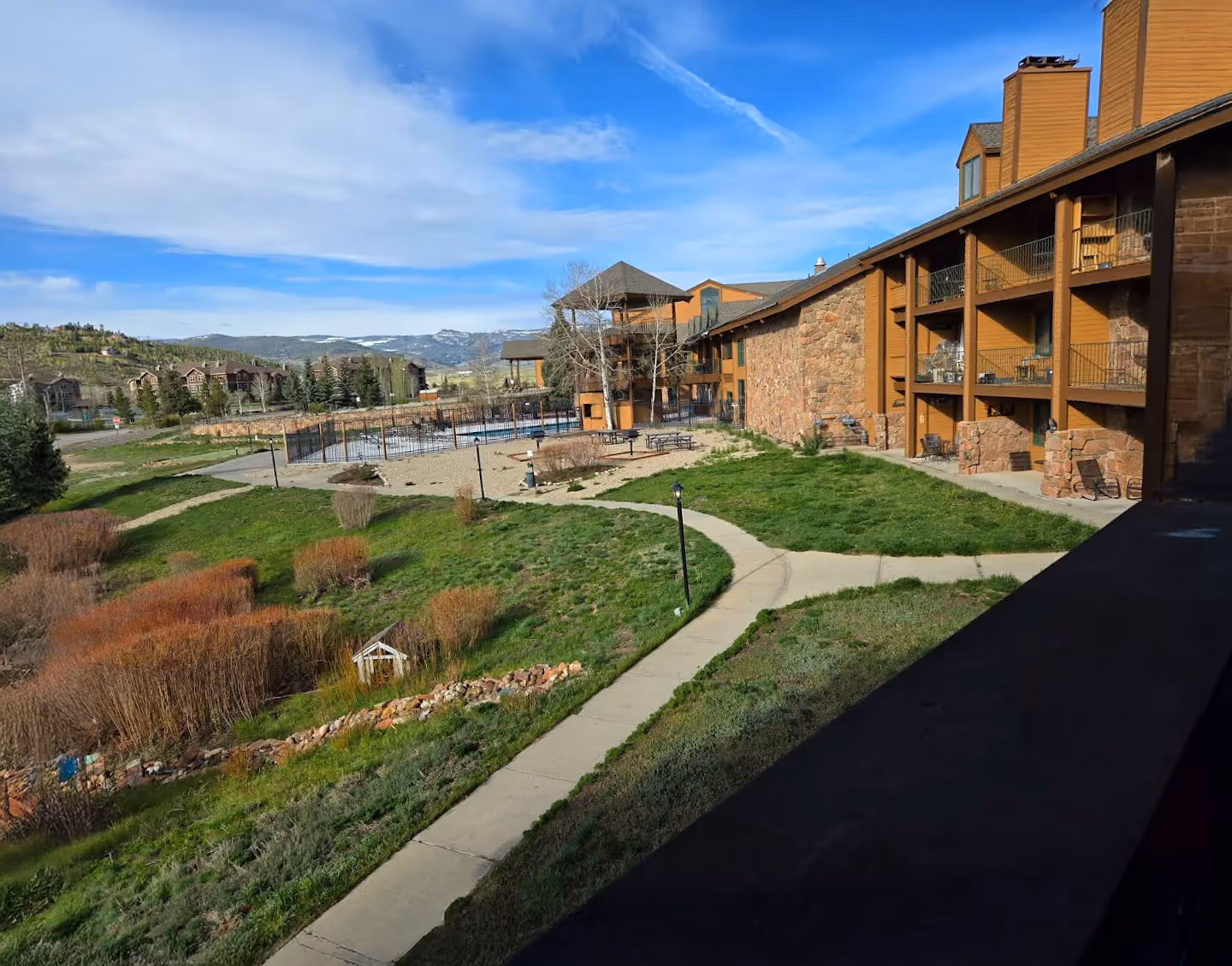 Mountain resort with a stone building, balconies, and pathways through green lawns on a sunny day. Mountains and blue sky create a serene backdrop.