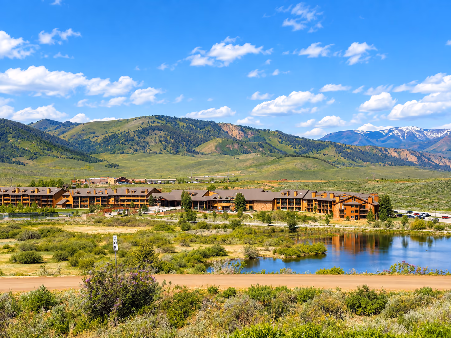 A sprawling lodge nestled in a lush, green valley surrounded by majestic mountains under a clear blue sky, with a tranquil pond in the foreground.