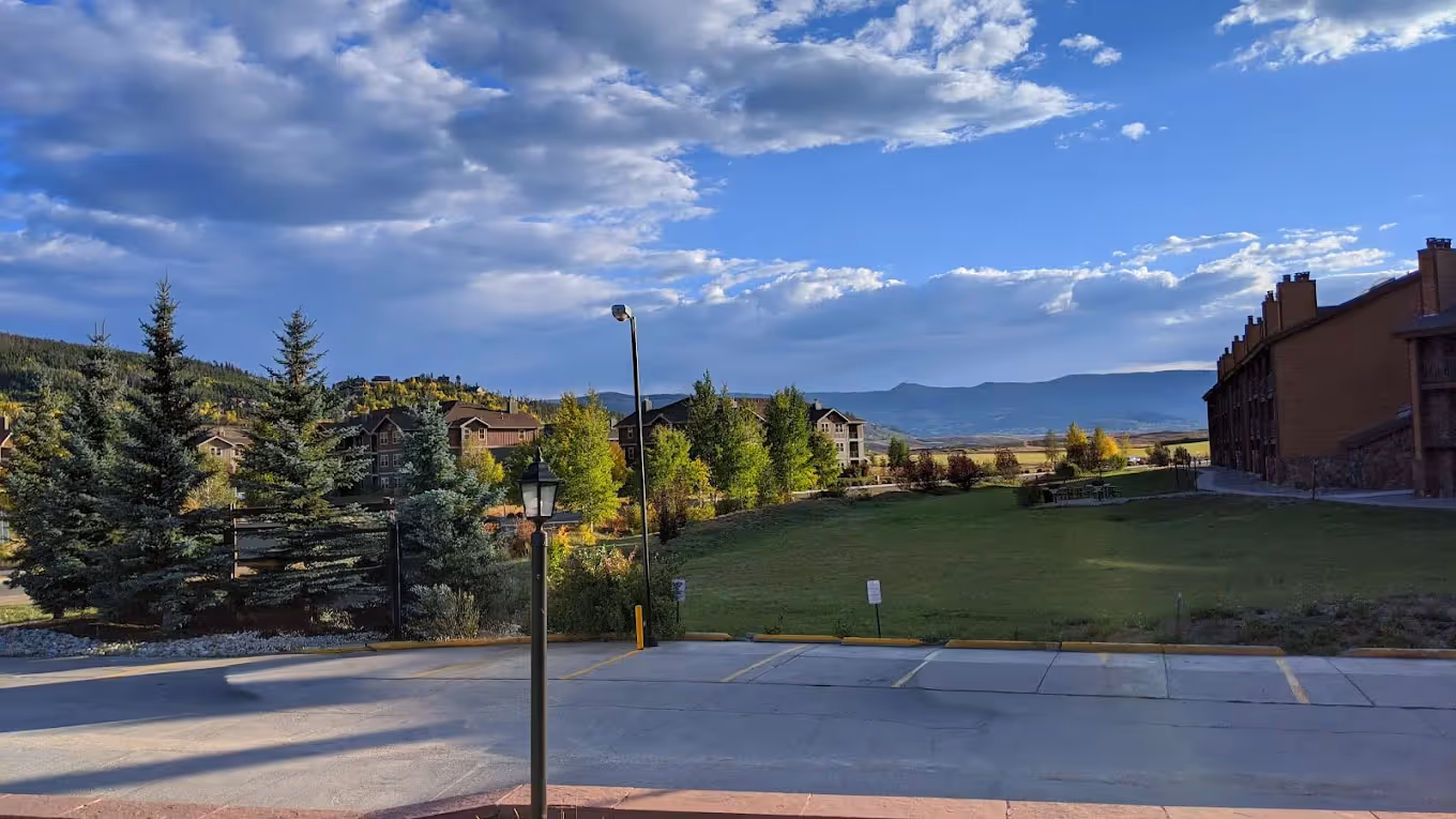 A serene landscape with a grassy field, rustic buildings, and evergreen trees under a partly cloudy sky. The scene conveys tranquility and openness.