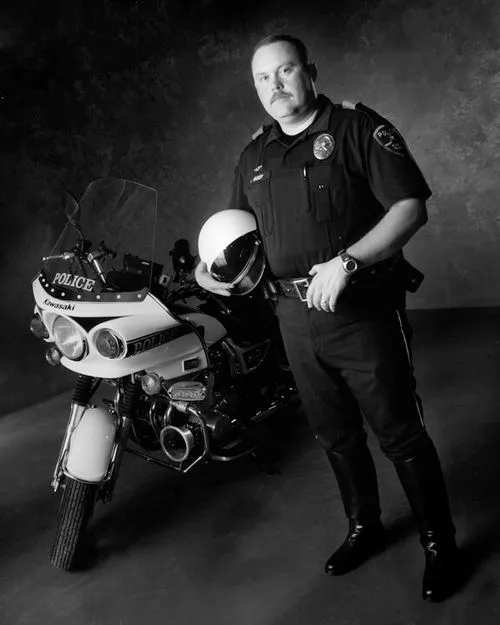 Black-and-white portrait of a police officer standing beside a Kawasaki police motorcycle, holding a helmet.
