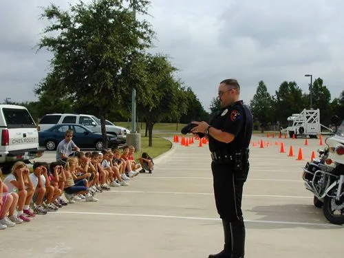 Police officer demonstrating firearm safety to a seated group of children in an outdoor parking lot.