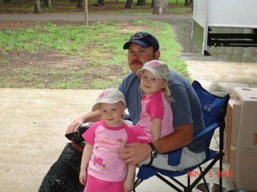 Man in a baseball cap sitting on a blue folding chair holding a girl in pink clothes, with another girl in pink standing beside them and a black dog nearby.