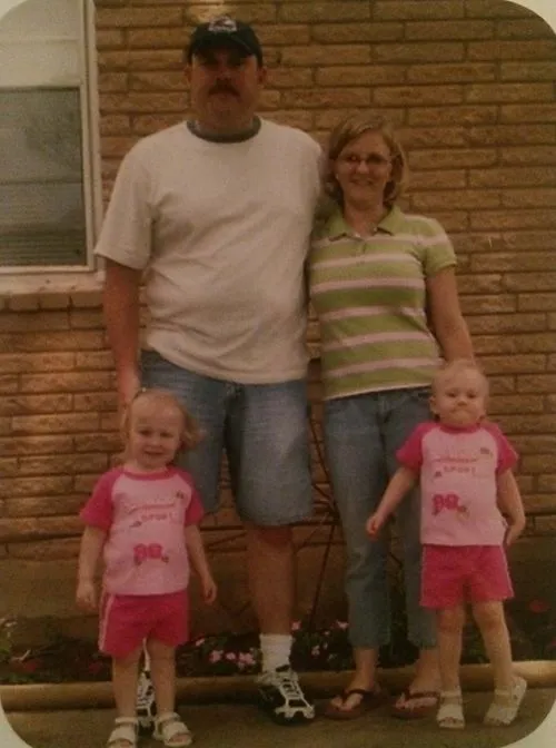 A family of four standing outside in front of a brick wall; a man in a white shirt and shorts, a woman in a striped polo and jeans, and two young girls in matching pink outfits.
