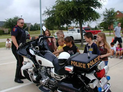 Police officer standing next to a police motorcycle while children gather around and look at it in an outdoor setting.