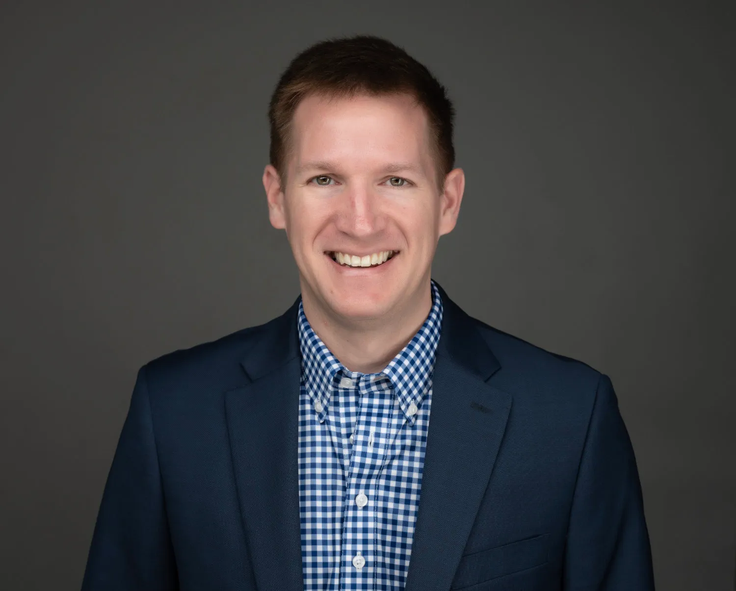 Smiling man wearing a navy blazer and blue checkered shirt against a gray background.