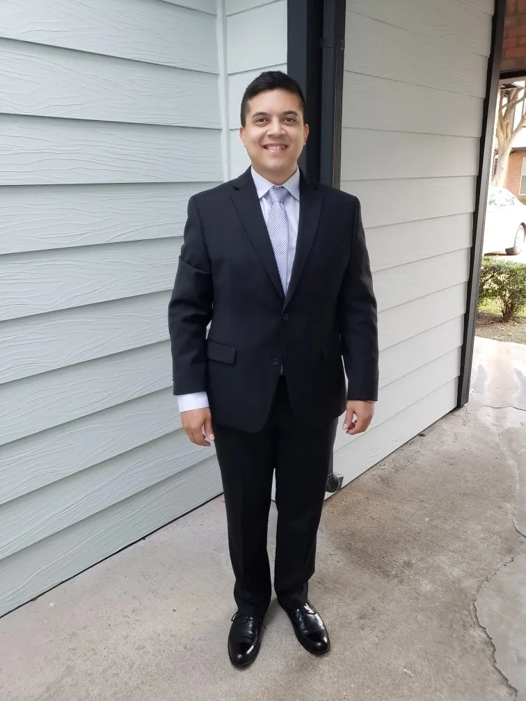 Man standing outdoors wearing a black suit, light blue shirt, patterned tie, and black dress shoes, smiling at the camera.