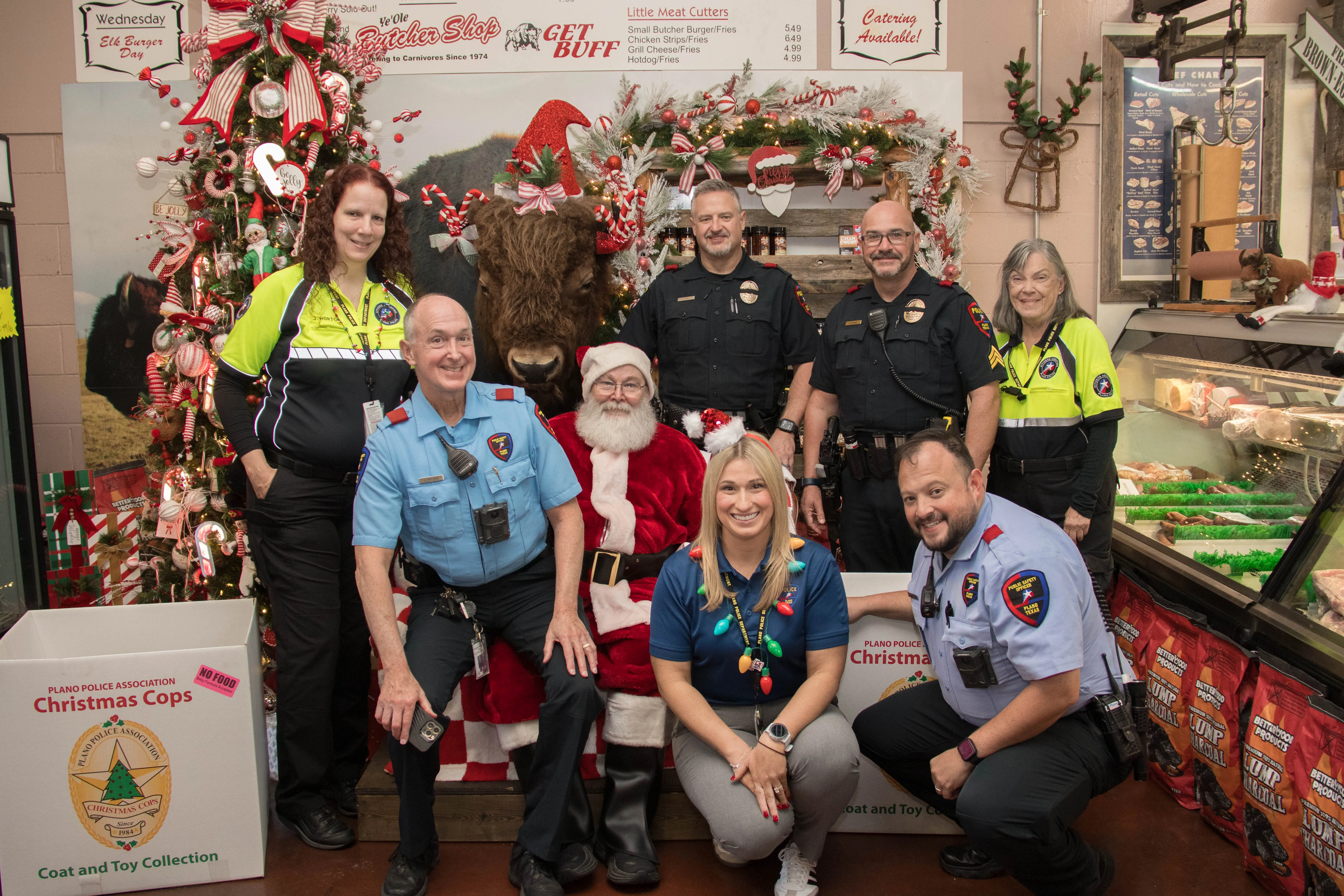 Group of police officers and volunteers posing with Santa Claus and a person in a buffalo costume in a festive Christmas decorated butcher shop.