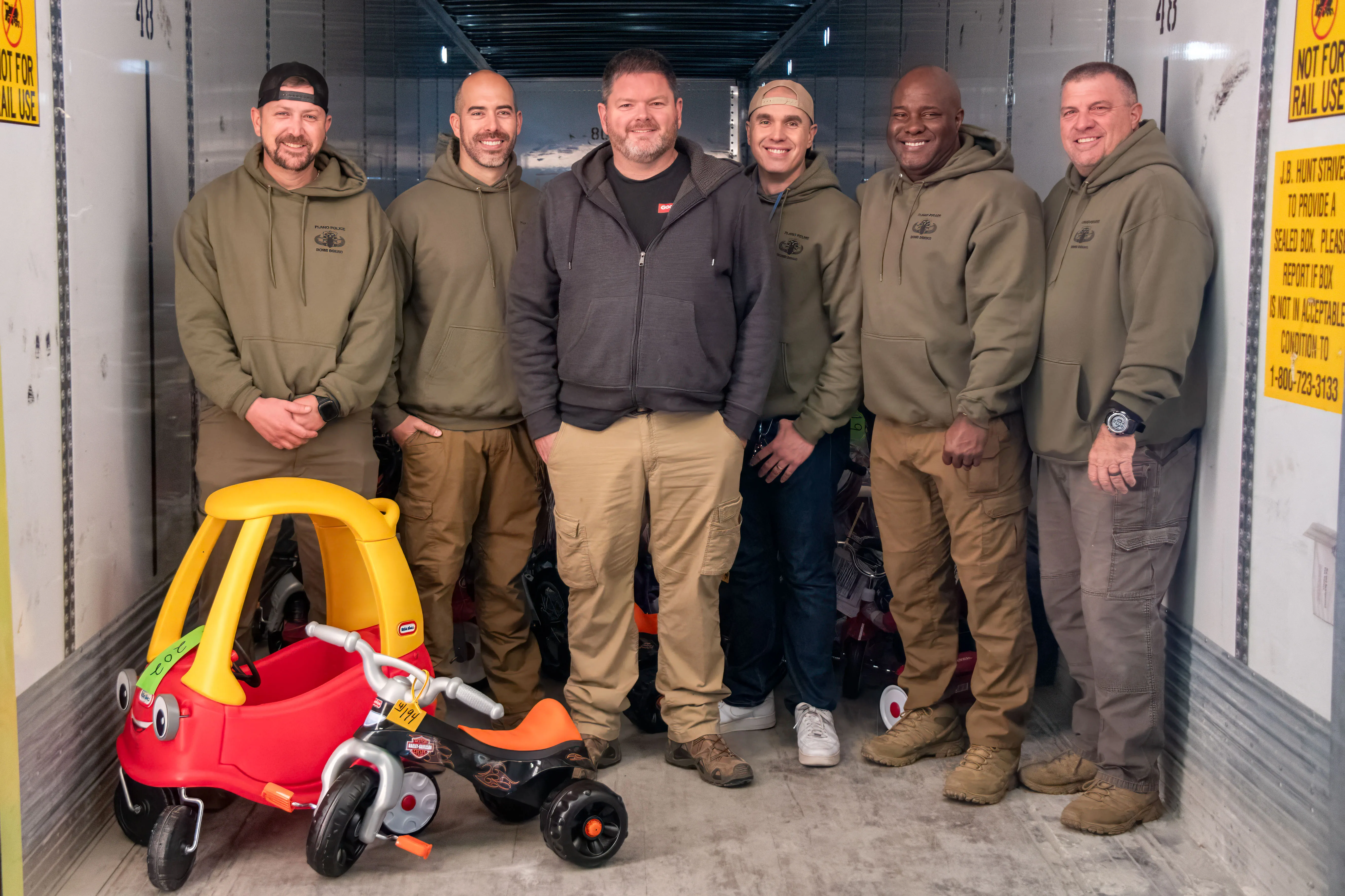 Six men, five in matching green hoodies and tan pants, standing inside a trailer with children's ride-on toy car and tricycle in front.
