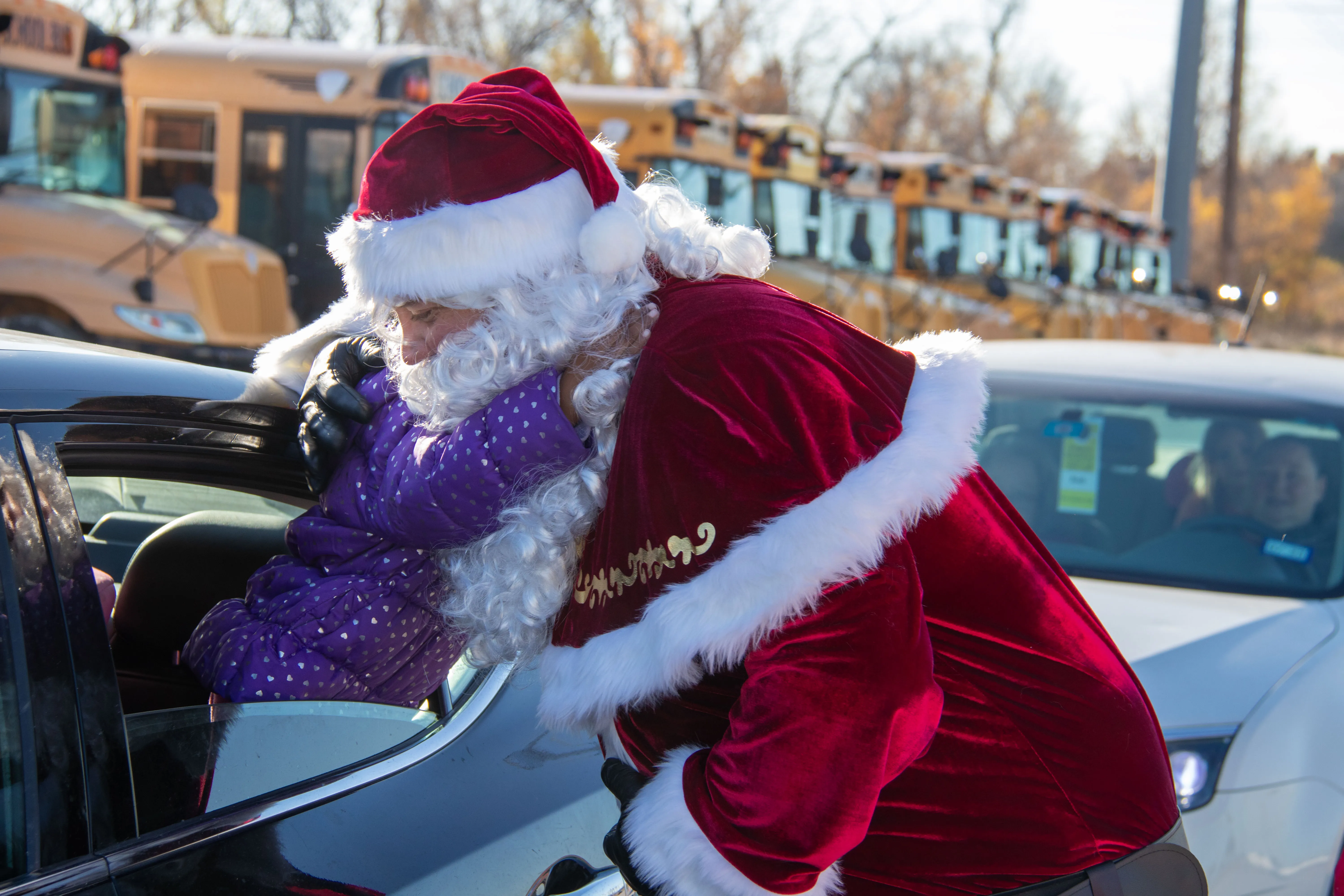 Santa Claus in red suit hugging a child in a car window with school buses in the background.