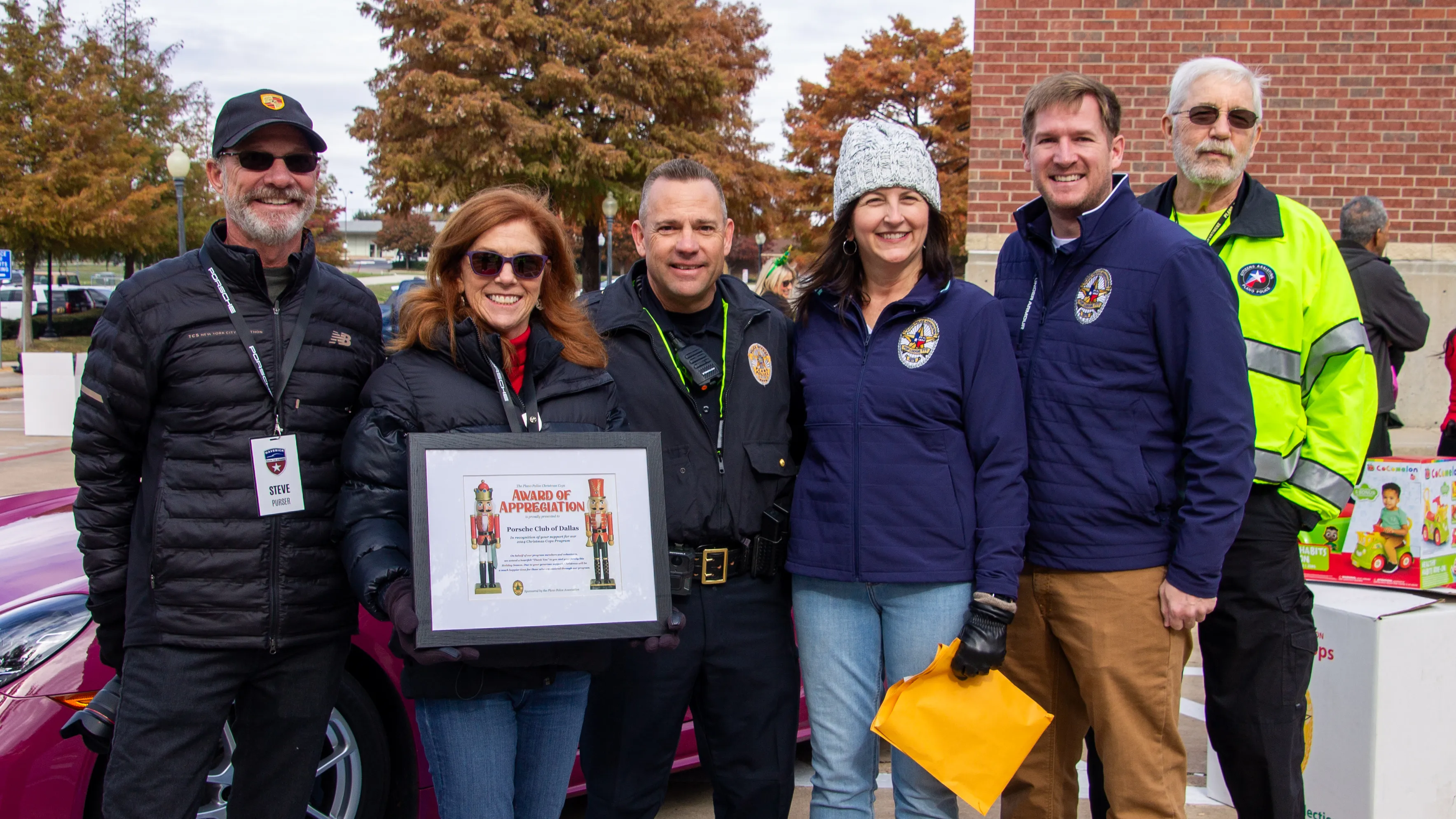 Group of six people, including police officers and event volunteers, standing outdoors with autumn trees in the background; one woman holds a framed award certificate.