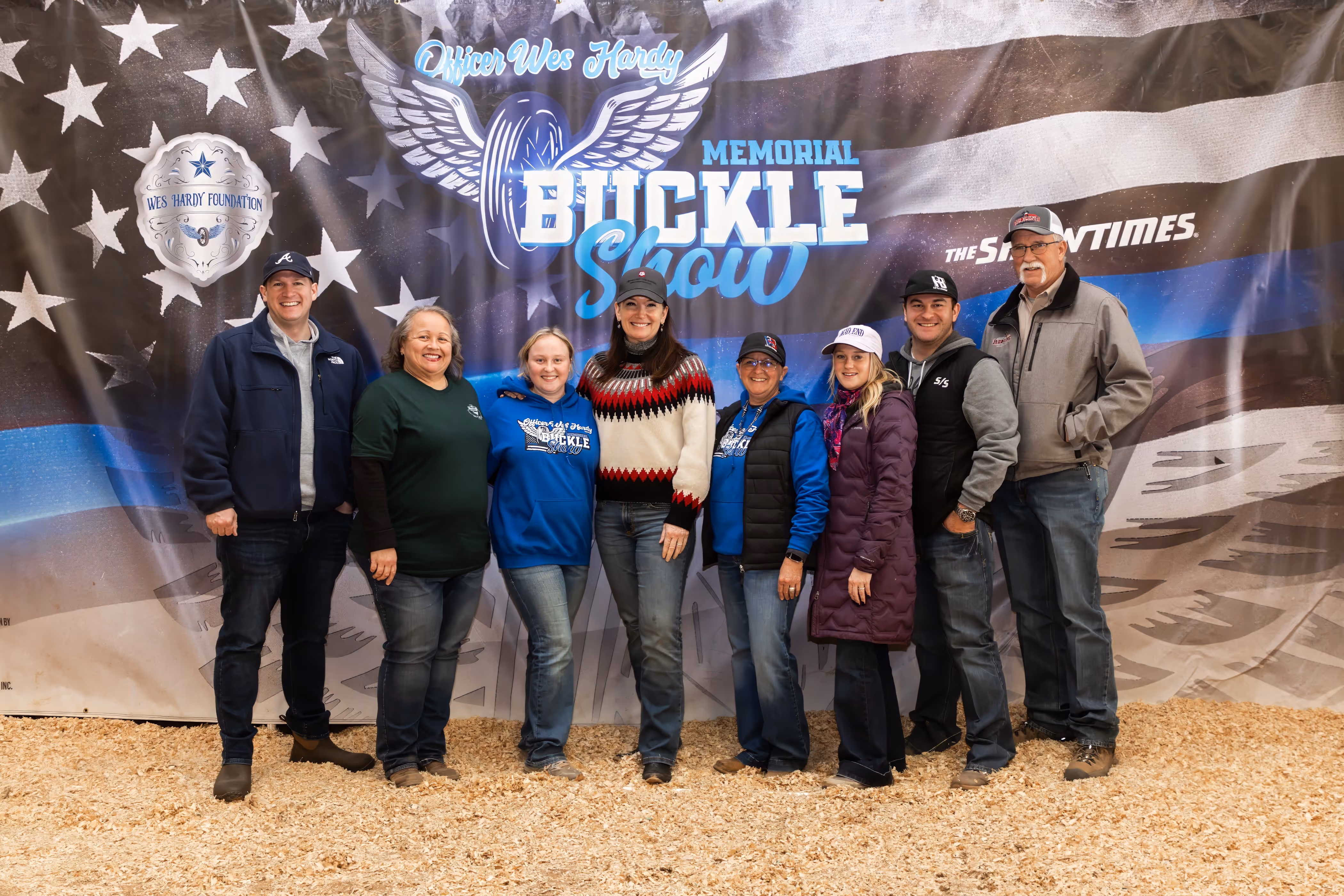 Eight people standing on wood chips in front of a banner for Officer Wes Hardy Memorial Buckle Show.
