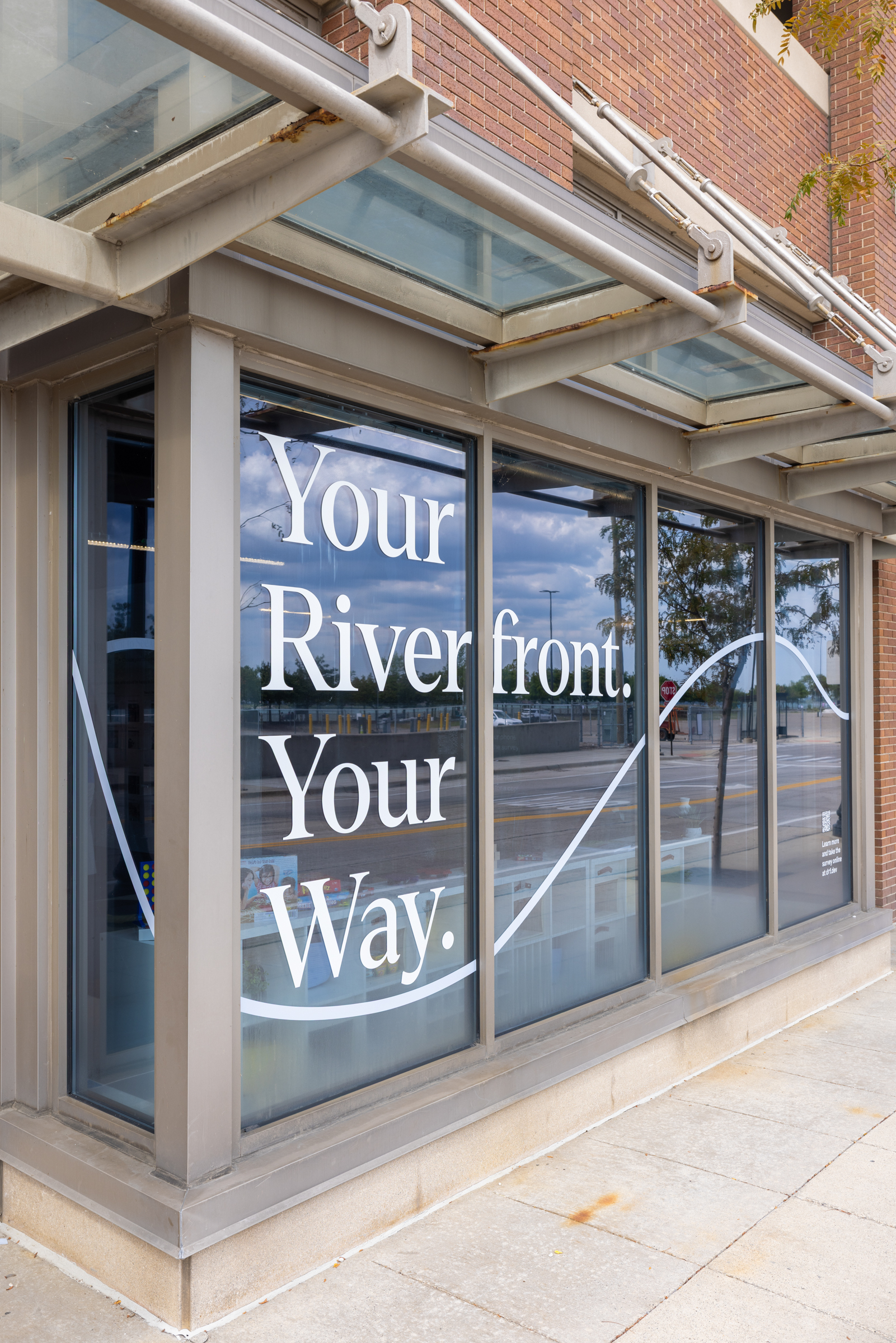 Exterior photo of the Community Hub at Beaubien Place in Detroit, Michigan, featuring window wrapping which says, "Your Riverfront. Your Way."