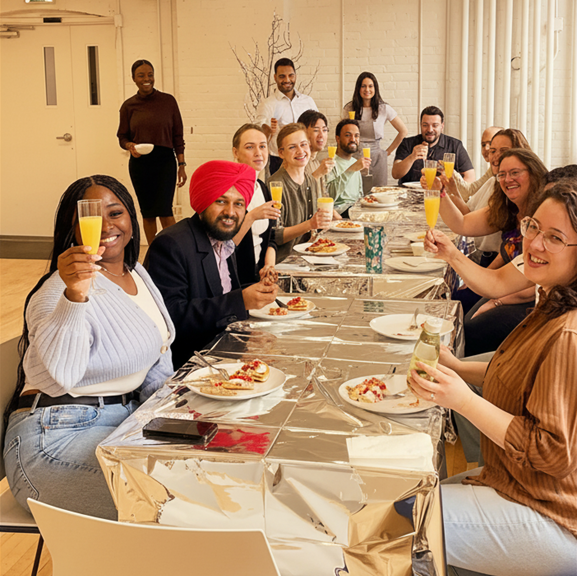 A diverse group of people sitting around a long table with food and drinks, raising glasses in a toast and smiling.
